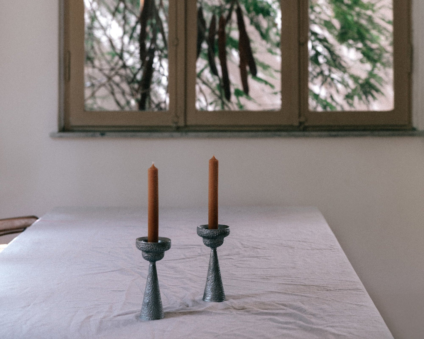 Two candles in green candle holders on a table with a window in the background showing trees.