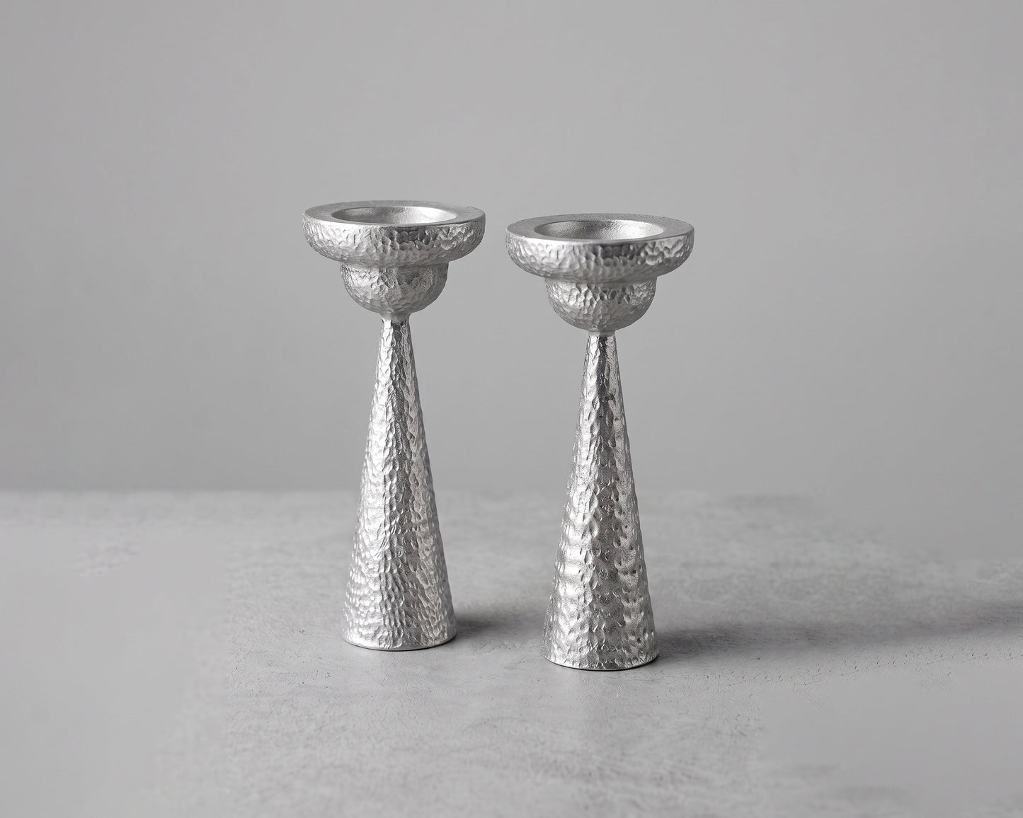 Two textured silver candlesticks standing upright on a light gray surface against a neutral gray background.