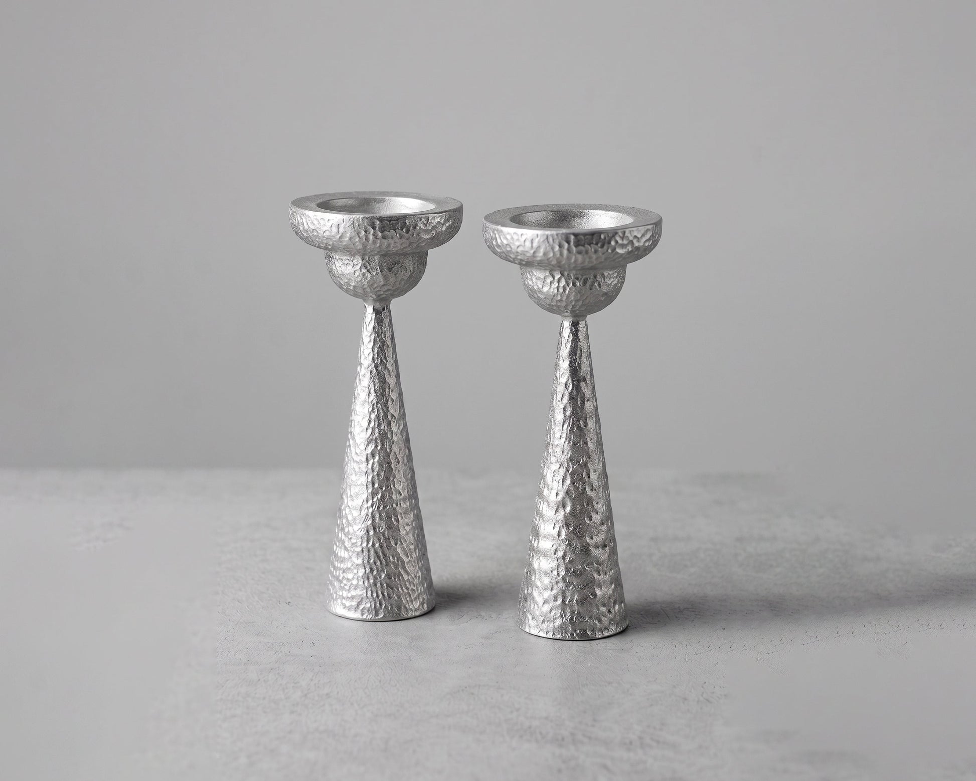 Two textured silver candlesticks standing upright on a light gray surface against a neutral gray background.