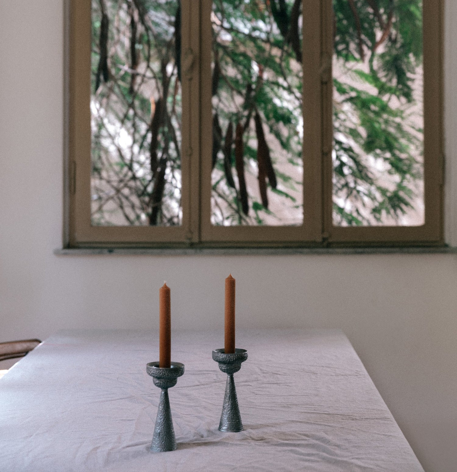 Two candles in green candle holders on a table with a window in the background showing trees.