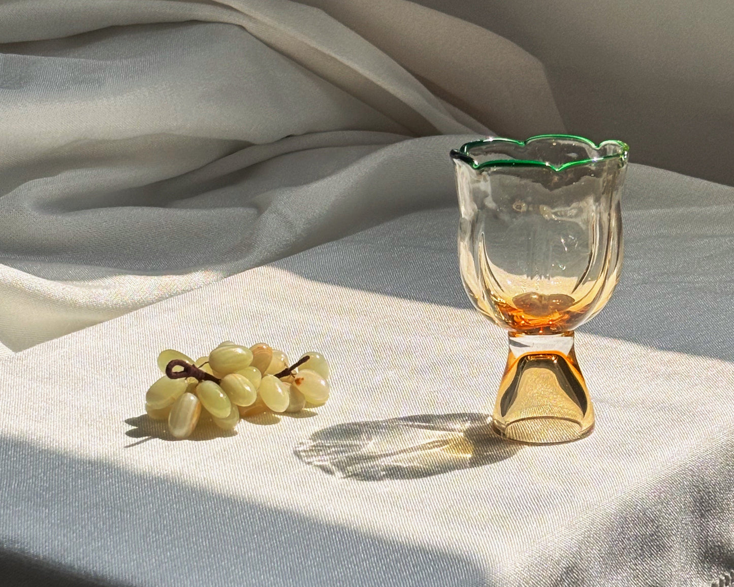 A clear glass goblet with a scalloped rim and amber-colored base, styled on a white tablecloth next to a small bunch of grapes in warm sunlight.