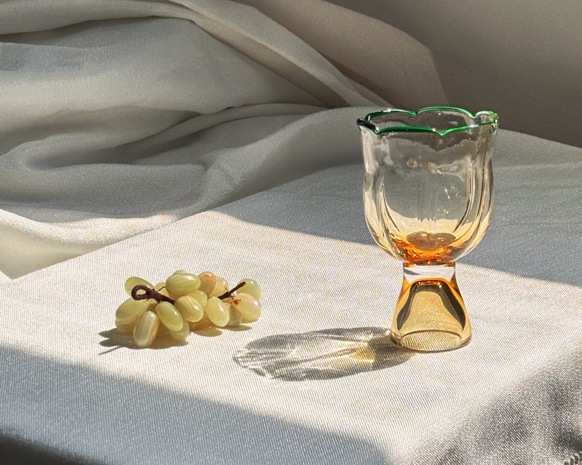 A clear glass goblet with a scalloped rim and amber-colored base, styled on a white tablecloth next to a small bunch of grapes in warm sunlight.