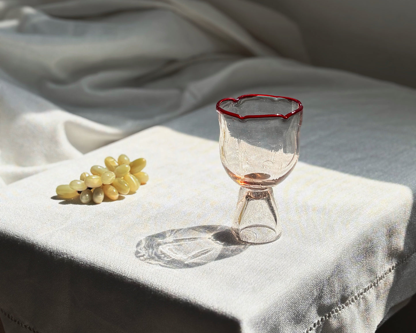 A clear glass goblet with a thin red rim sits on a white embroidered tablecloth next to a small bunch of pale grapes in a softly lit interior setting.