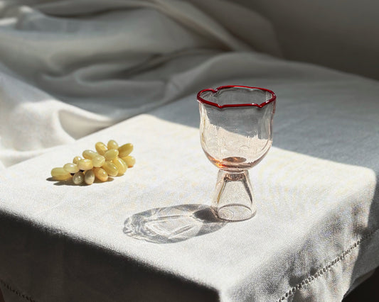 A clear glass goblet with a thin red rim sits on a white embroidered tablecloth next to a small bunch of pale grapes in a softly lit interior setting.